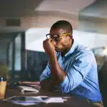 A man sits at his desk holding nose while dealing with a headache - How To Avoid Vyvanse Headaches